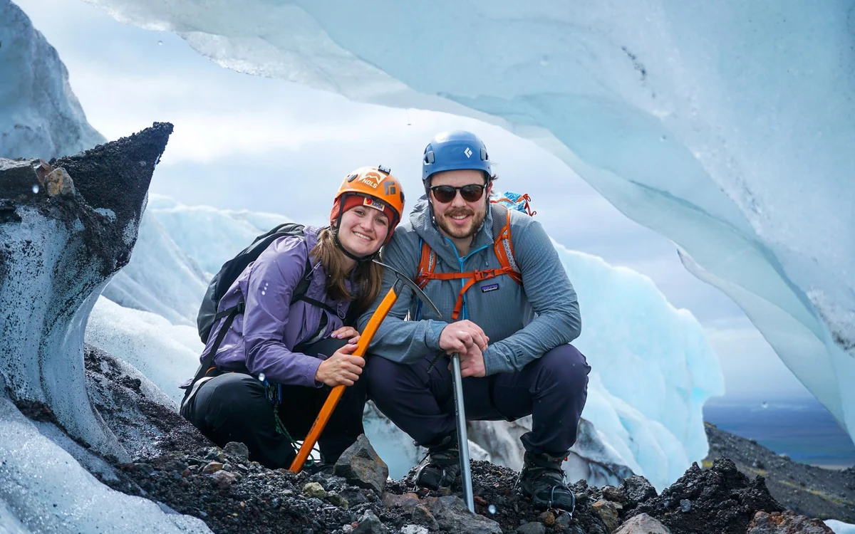 Zwei Wanderer mit Helmen hocken mit Eispickeln auf dem Gletscher und lächeln in die Kamera
