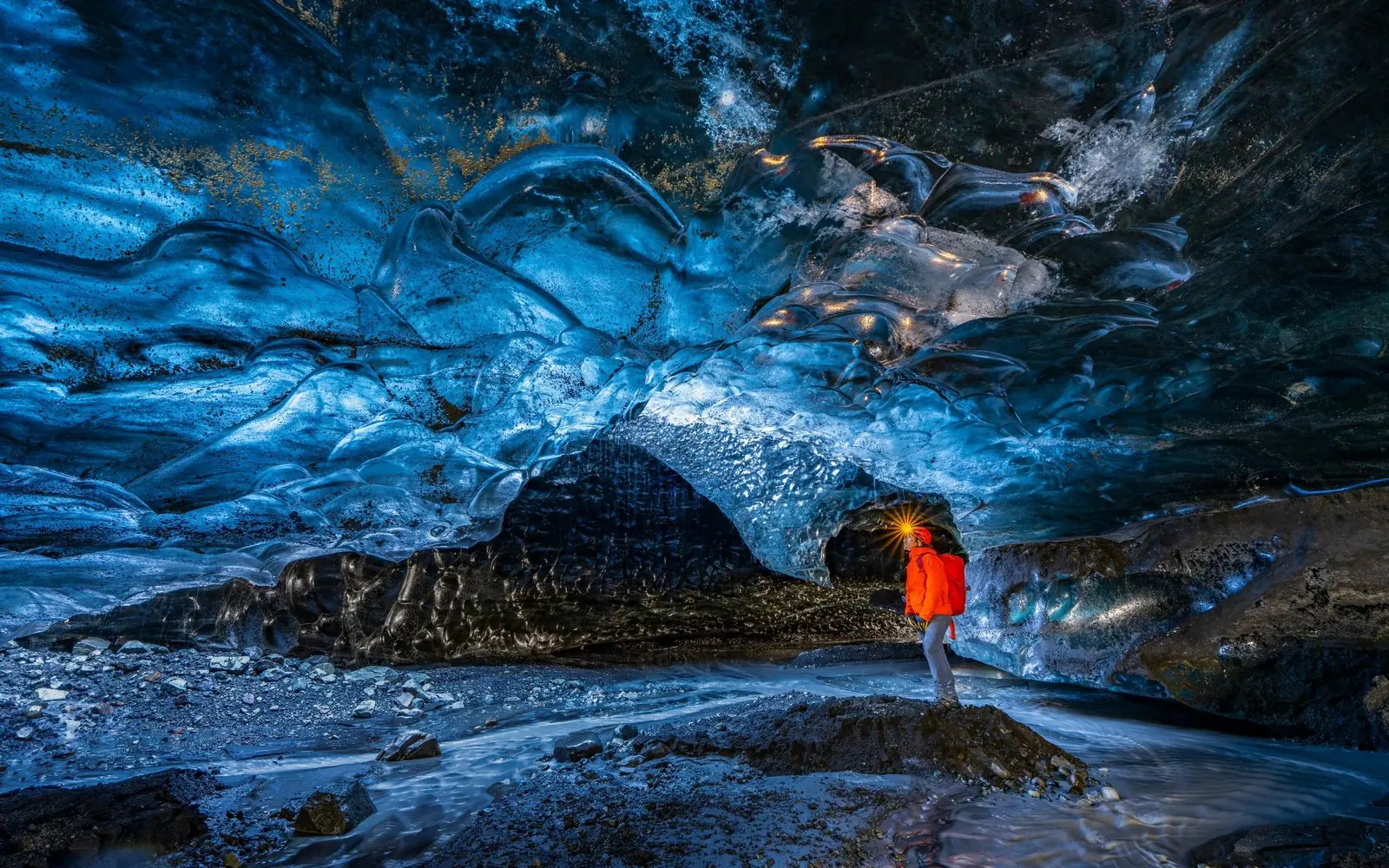 Eine Wanderin in roter Jacke steht unter dem blauen Eisgewölbe einer Gletscherhöhle, Schmelzwasser läuft über den dunklen Boden