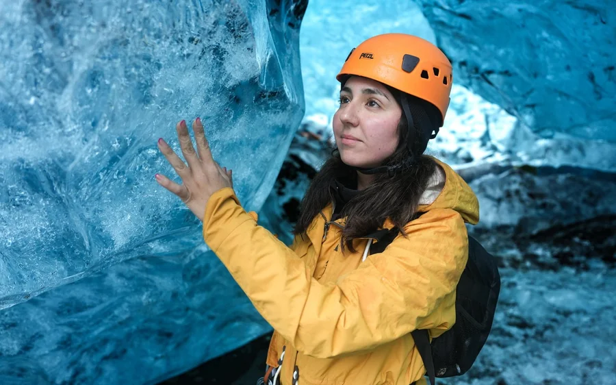 Eine Frau in gelber Jacke und orangefarbenem Helm steht mit erhobenen Händen in einer blauen Eishöhle
