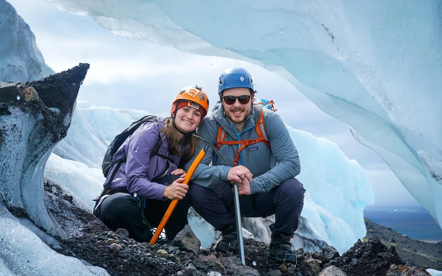 Zwei Wanderer mit Helmen und Eisäxten kauern auf dem Gletscher und lächeln in die Kamera
